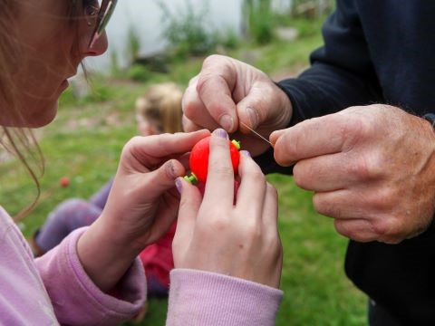 four hands are in the picture, two around a bright orange bobber and two holding the fishing line to assist in attaching the bobber to the line.