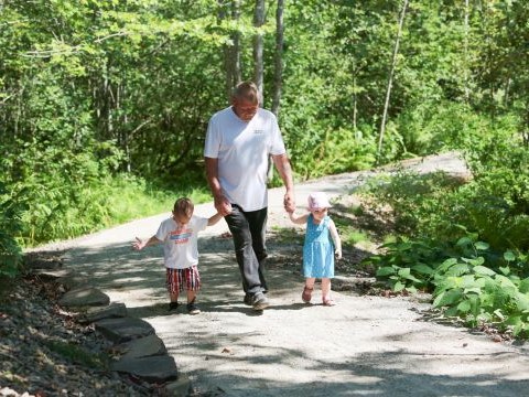 Two young children holding an adult’s hands and walking down a path in the woods on a sunny day.