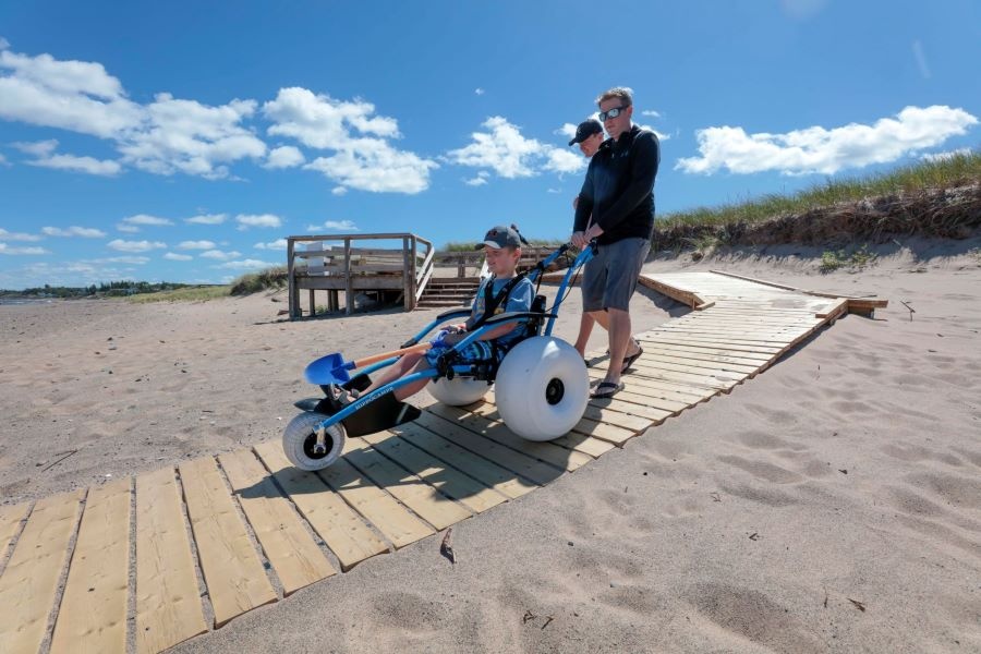 A boy using a Hippocampe wheelchair with his family on a beach boardwalk.